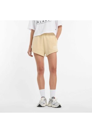Person trägt beige Shorts mit Kordelzug, weiße Kniestrümpfe, graue und weiße Turnschuhe und ein kurzes weißes Shirt mit schwarzem Text.
