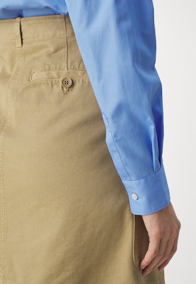 Camicia azzurro chiaro con maniche lunghe, abbinata a pantaloni di stoffa beige con una tasca posteriore e dettaglio con bottone.