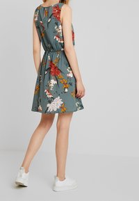 Kwiatowa krótka sukienka w kolorze teal z czerwonymi, różowymi i pomarańczowymi kwiatami. Posiada dopasowaną talię i rozkloszowany dół, zestawiona z białymi trampkami.