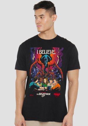 Young man wearing black Stranger Things t-shirt featuring characters, large spider-like creature, and text "I Believe" and "The Right Side Up."