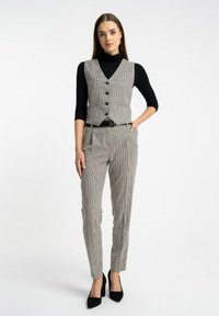 Kobieta stoi z jedną ręką w kieszeni, ubrana w kamizelkę w kratę oraz dopasowane spodnie, pod które ma na sobie czarny dopasowany top i czarne szpilki, na białym tle.