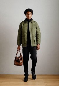 Quiltet olivengrøn jakke med to forlommer, båret over en sort turtleneck, parret med sorte bukser og holder en brun lædertaske.