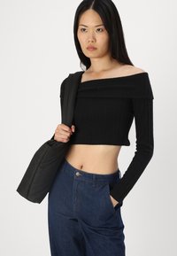 Černý žebrovaný crop top bez ramínek s dlouhými rukávy, kombinovaný s tmavě modrými džíny s vysokým pasem. Modelka drží černou kabelku přes rameno.