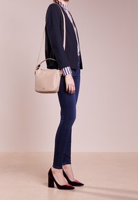 Žena oblečená v námořnickém blejzru, pruhované košili, tmavých skinny džínách, bordó sametových lodičkách na vysokém podpatku a béžové crossbody tašce, stojící bokem.