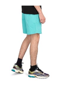 Shorts turquoise en tissu coton, avec une coupe droite et des poches latérales. Associés à des chaussures de sport multicolores. Chaussettes noires visibles.