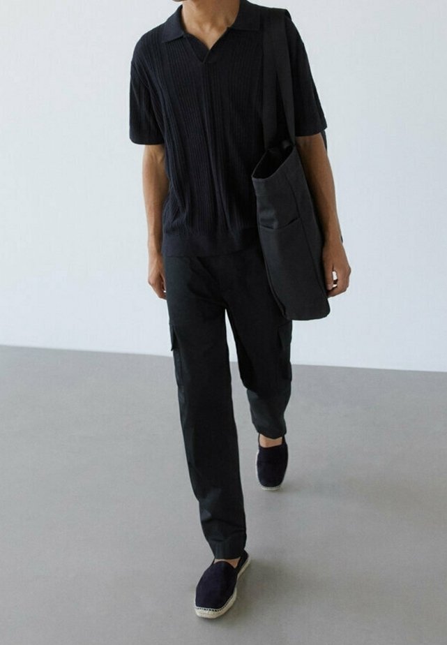 Personne portant un t-shirt côtelé noir à manches courtes, un pantalon cargo noir, des chaussures noires sans lacets et portant un grand sac noir à l'épaule.