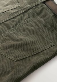 Grüner Cordstoff mit einer aufgesetzten Tasche und vertikaler gerippter Struktur. Das Material ist langlebig und hat einen dezenten Glanz.