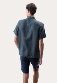 Dunkelgrünes Leinenhemd mit kurzen Ärmeln und Kragen, strukturierter Stoff und gerader Schnitt, kombiniert mit marineblauen Shorts. Ansicht von hinten.