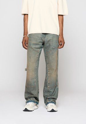 Mann trägt verblasste blaue Jeans mit lockerem Sitz und Kordelzug an den Säumen, weiße Turnschuhe und ein cremefarbenes kurzärmeliges Oberteil, steht vor einem schlichten Hintergrund.