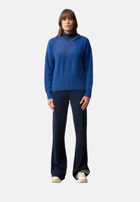 Blå stickad huvtröja med dragkedja fram och huva, tillsammans med mörkblå vida byxor. Modellen bär ljusa sneakers.