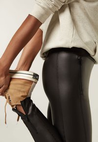 Leggings neri e lucidi con dettagli a zip, abbinati a una sneaker beige tenuta in mano, su uno sfondo neutro.