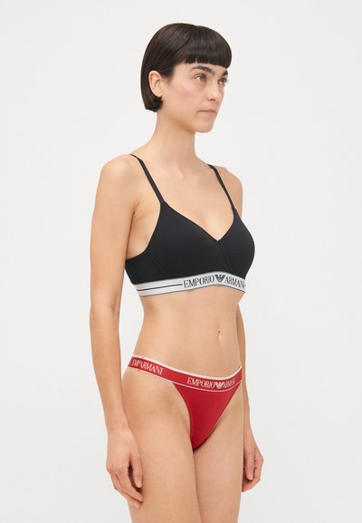Black bralette with thin straps and white logo band, paired with red thong featuring logo waistband. Smooth fabric, minimalistic design.