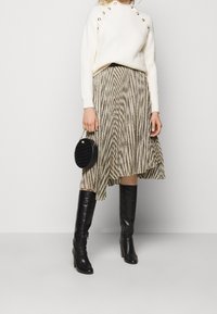 Žena v bílé svetr s knoflíkovým detailem, přiléhavé midi sukni v hnědých a béžových pruzích, černých kožených vysokých botách a držící kulatou černou kabelku.