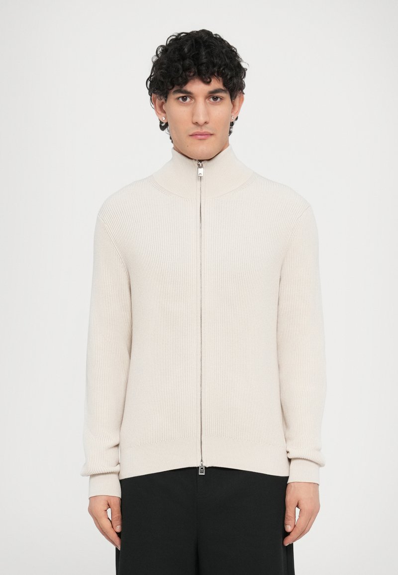 Μπεζ ριμπ πλεκτό πουλόβερ με φερμουάρ και ψηλό γιακά, με ασημένιο φερμουάρ και μακριά μανίκια, συνδυασμένο με σκούρο παντελόνι.