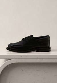 Chaussure habillée noire en cuir à lacets posée sur une surface en pierre à bords arrondis avec un fond beige neutre.