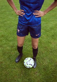 Blue athletic jersey with textured pattern, paired with dark shorts featuring a rooster logo. Black socks and cleats; a soccer ball rests on grass.
