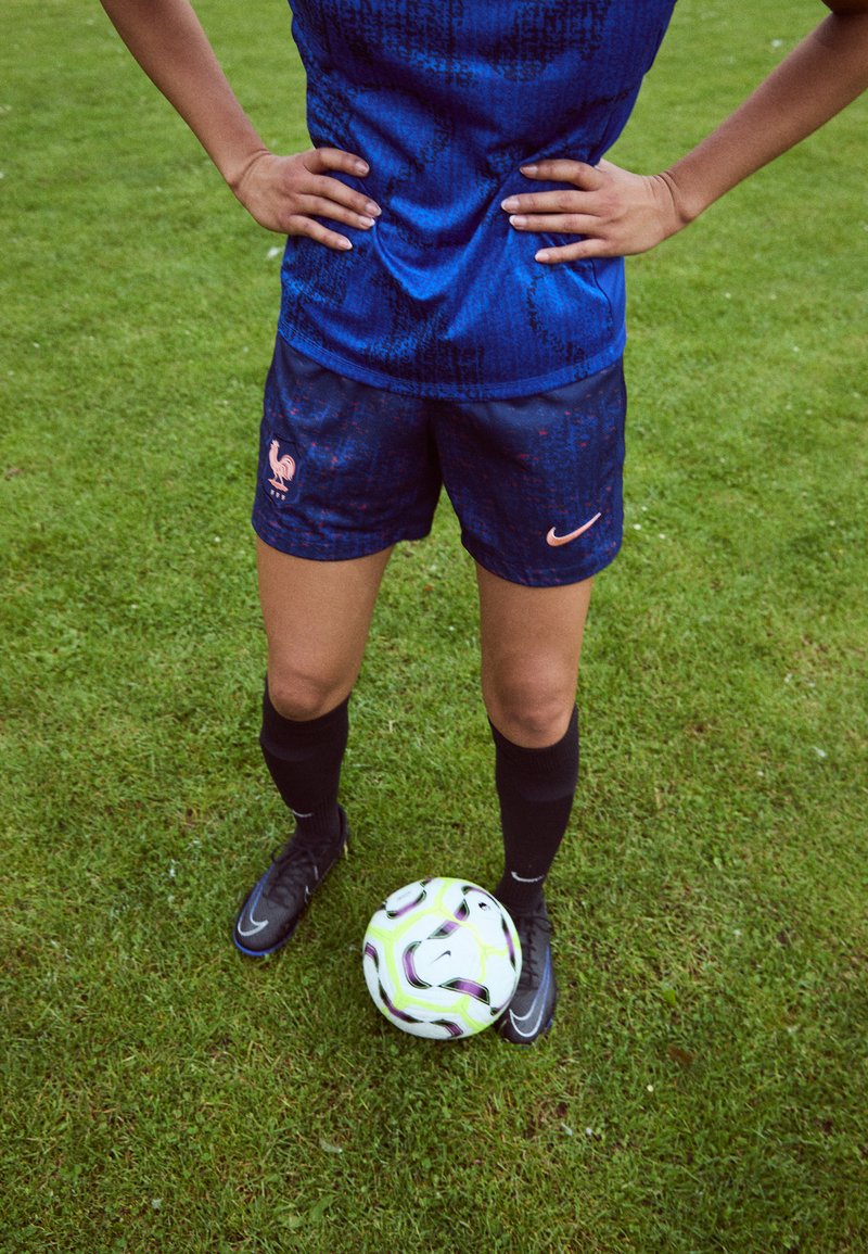 Blue athletic jersey with textured pattern, paired with dark shorts featuring a rooster logo. Black socks and cleats; a soccer ball rests on grass.