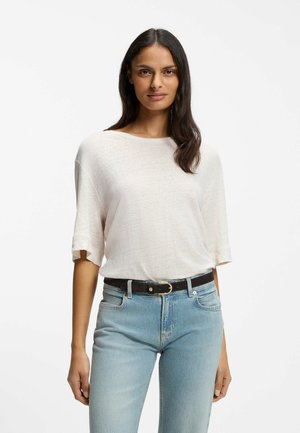 Frau mit langen dunklen Haaren, die ein lockeres weißes kurzärmeliges Oberteil trägt, das in eine hellblaue Jeans mit schwarzem Gürtel gesteckt ist, vor weißem Hintergrund.