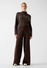 Braunes passendes Set mit einem kurz geschnittenen Top mit hohem Kragen und weit geschnittenen Hosen. Weicher Stoff mit glatter Textur, dezentes Logo-Detail am Top.