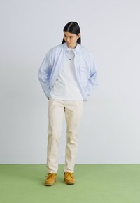 Cămașă albastră deschis, cu butoni, peste un tricou alb, purtată cu pantaloni bej și pantofi galbeni cu șireturi, pe un podea verde.
