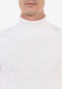 Pull en maille blanche avec col roulé, présentant un motif en grille, un col côtelé et une texture douce. Le design possède des détails subtils sur l'ensemble.