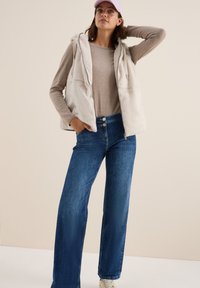 Ljusbeige fuskpälsvest med huva över en tan långärmad topp, kombinerad med blå vida jeans och vita sneakers. Lavendelfärgad keps ger en extra detalj.