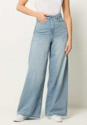 Kvinde iført lyseblå, højtaljede viddeben jeans, hvide sneakers og en hvid ærmeløs top, med den ene hånd i lommen.