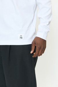 Mörkhyad person som bär en vit långärmad tröja med etiketten "M 73" nära fållen och svarta byxor, handen avslappnad vid sidan.