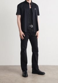 Zwarte short-sleeve overhemd met knoopjes en een kraag, subtiel logo, gecombineerd met zwarte jeans en zwarte laarzen, wat een monochrome uitstraling creëert.