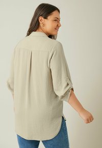 Helles beige Hemd mit leicht strukturierter Oberfläche, geknöpften Ärmel, Rückenschlitz und abgerundetem Saum, kombiniert mit blauen Jeans.