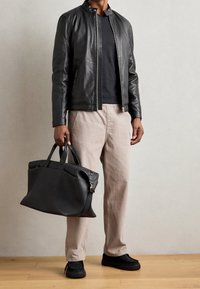 Schwarze Lederjacke mit Reißverschluss und Stehkragen, kombiniert mit beigen Hosen und einer schwarzen strukturierten Tragetasche. Schwarze Slippers.