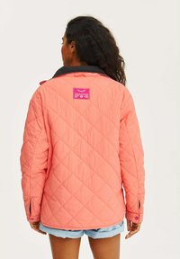 Přikrývková bunda v jasně korálové barvě s černým límcem, s růžovou nálepkou na zadní straně a s patentkami na rukávech.