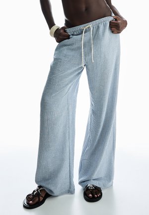 Person wearing loose, light blue and white pinstriped pants with drawstring, black sandals, and a white bracelet, hands in pockets.