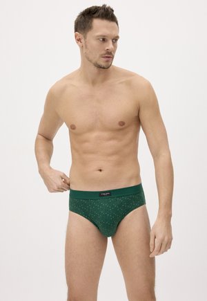 Hombre sin camiseta con cabello corto, vistiendo calzoncillos verdes con estampado, de pie sobre un fondo blanco liso, mirando hacia su izquierda.