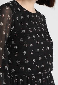 Svart blus med genomskinliga långa ärmar, med ett blommönster i vitt och rött. Slät textur och rund halsringning.