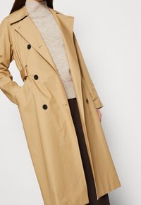 Gabardina beige con un diseño de doble botonadura, botones negros, cuello oversized y bolsillos laterales, llevada sobre un suéter neutro.