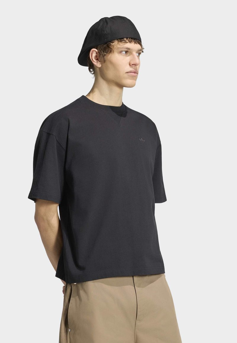 Jeune homme portant un t-shirt noir oversize, une casquette noire à l'envers et un pantalon beige, debout devant un fond clair uni.