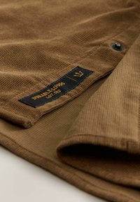 Brown corduroy fabric with a ribbed texture. Features a black label with gold text and a button accent near the edge.