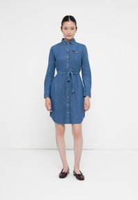 Kvinne står iført en blå denimskjortekjole i kneelengde med lange ermer, knappestilling foran, knyttet livbelte og burgunder ballet-sko.