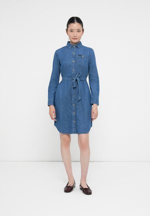 Kvinna som står klädd i en knälång blå denimskjortklänning med långa ärmar, knäppning fram, knytbart midjeband och bordeauxfärgade balettklackar.