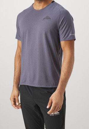 Homme portant un t-shirt violet Nike Trail à manches courtes et un pantalon noir Nike Trail, debout sur un fond clair uni.