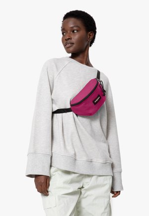Fanny packki, joka on kirkasta pinkkiä kangasta ja jossa on musta vetoketju ja hihna, käytetään harmaan collegepuseron ja vaaleiden cargo-housujen päällä.