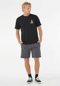 Sort t-shirt med korte ærmer og et guld-logo, parret med grå shorts med sidelommer og sorte sneakers med hvide detaljer.