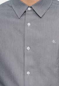 Ανδρικό πουκάμισο με κάθετες γκρι και λευκές ρίγες, κουμπιά μπροστά, με μυτερό γιακά και διακριτικό λογότυπο "CK" στο αριστερό μέρος του στήθους.