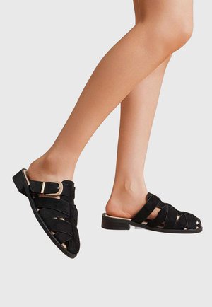 Jambes portant des sandales plates ouvertes en suède noir avec plusieurs lanières et une boucle latérale sur un fond gris clair.