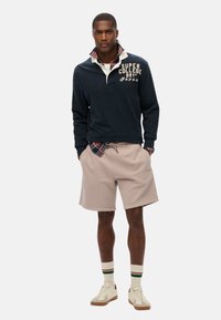 Pull noir marine avec un col blanc, texte "SUPER COLLEGE", shorts beiges, baskets crème et chaussettes rayées. Chemise à carreaux visible en dessous.