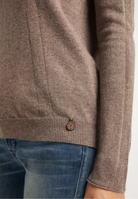 Close-up van een persoon die een bruine gebreide trui draagt met een klein rond ankerlogo nabij de onderrand en blauwe spijkerbroek.