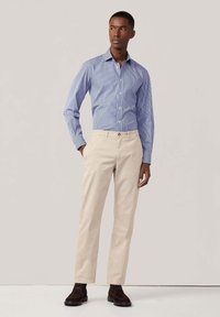 Camisa azul claro a rayas con botones y mangas largas, pantalones crema y botas de ante marrón oscuro hasta el tobillo. El modelo se encuentra de pie con las manos en los bolsillos.