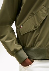 Persoon in een olijfgroene jas met klepzak en elastische manchetten, hand in de zak van witte broek.