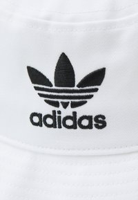 Bela kapa s ploskim vizirjem, izdelana iz blaga, s črnim vezenim logotipom adidas tridelnim oblikovanjem in besedilom. Teksturirana površina.